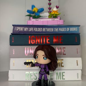 Pu&ograve; includere: Una pila di libri con i titoli "I Spent My Life Folded Between the Pages of Books", "Ignite Me", "Unite Me" e "Restore Me". Una statuina di una donna in tuta viola che tiene una pistola &egrave; di fronte ai libri. Fiori decorativi in cima.