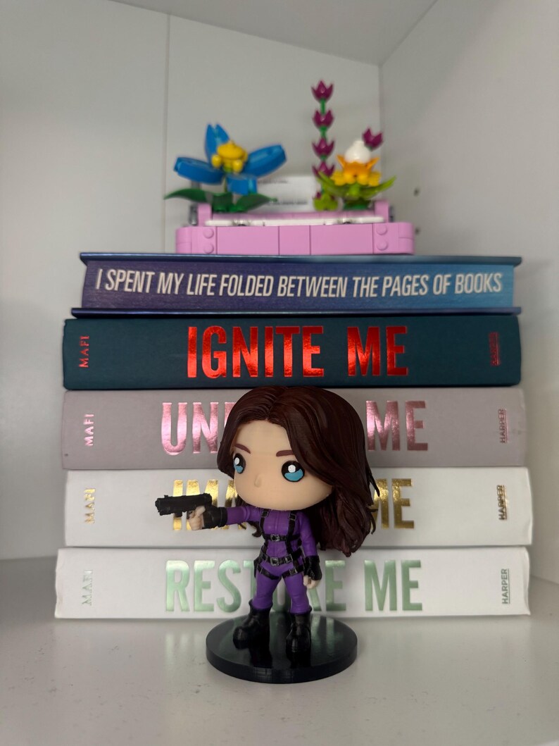 Pu&ograve; includere: Una pila di libri con i titoli "I Spent My Life Folded Between the Pages of Books", "Ignite Me", "Unite Me" e "Restore Me". Una statuina di una donna in tuta viola che tiene una pistola &egrave; di fronte ai libri. Fiori decorativi in cima.