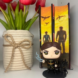 May include: A Funko Pop figure of a character with brown hair, holding a blue lightning bolt, and a black dragon figurine. The figure is on a black base, next to two books and a vase of red tulips.