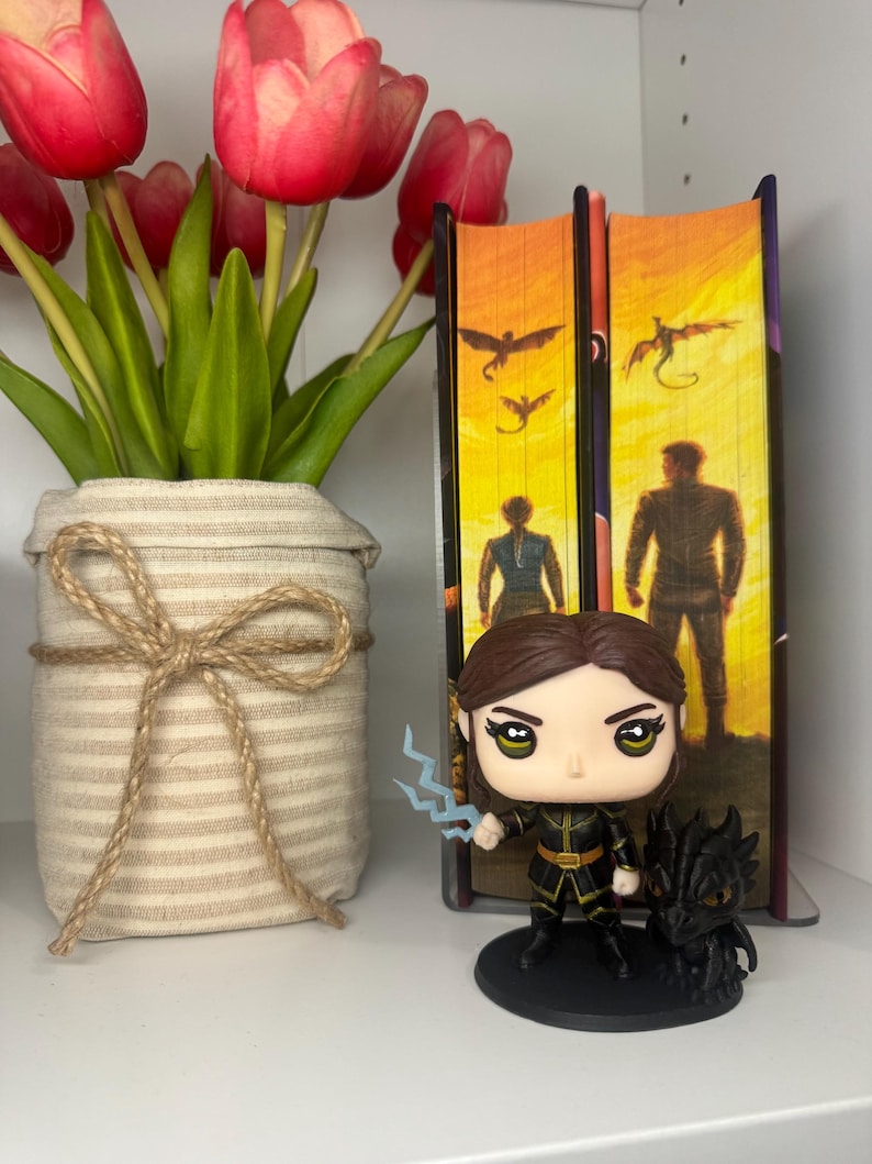 May include: A Funko Pop figure of a character with brown hair, holding a blue lightning bolt, and a black dragon figurine. The figure is on a black base, next to two books and a vase of red tulips.