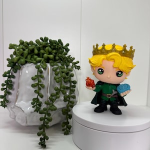 May include: A figurine of a character with blonde hair and a gold crown, wearing a green outfit, holding a flame and a blue orb. The figurine is on a white platform, with a white ceramic planter and green trailing plant in the background.
