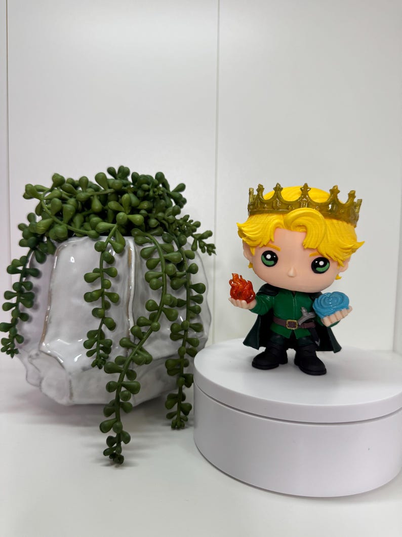 May include: A figurine of a character with blonde hair and a gold crown, wearing a green outfit, holding a flame and a blue orb. The figurine is on a white platform, with a white ceramic planter and green trailing plant in the background.