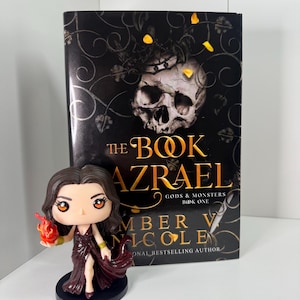 May include: A book titled "The Book Azrael" with a skull graphic and a figurine of a woman in a burgundy dress holding a flame. The book is part of the "Gods & Monsters" series by Amber V. Nicole.