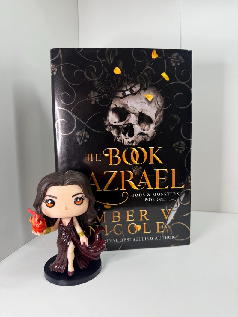 May include: A book titled "The Book Azrael" with a skull graphic and a figurine of a woman in a burgundy dress holding a flame. The book is part of the "Gods & Monsters" series by Amber V. Nicole.
