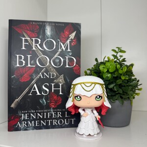 May include: A book titled "From Blood and Ash" by Jennifer L. Armentrout, with a Funko Pop figure of a character from the book. The book cover is black with red accents and a silver sword. The figure is white with red hair and a gold headpiece.