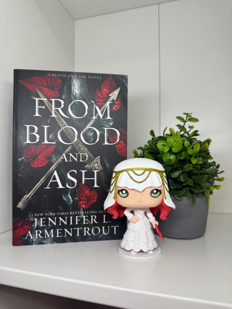 May include: A book titled "From Blood and Ash" by Jennifer L. Armentrout, with a Funko Pop figure of a character from the book. The book cover is black with red accents and a silver sword. The figure is white with red hair and a gold headpiece.