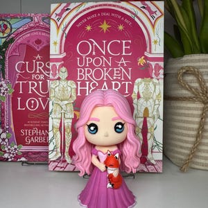 K&ouml;nnte beinhalten: Eine rosa Prinzessinnenfigur, die einen kleinen orangefarbenen Fuchs h&auml;lt, vor einem Buch mit dem Titel "Once Upon a Broken Heart". Die Figur hat gro&szlig;e blaue Augen und rosa Haare. Das Buchcover ist rosa.