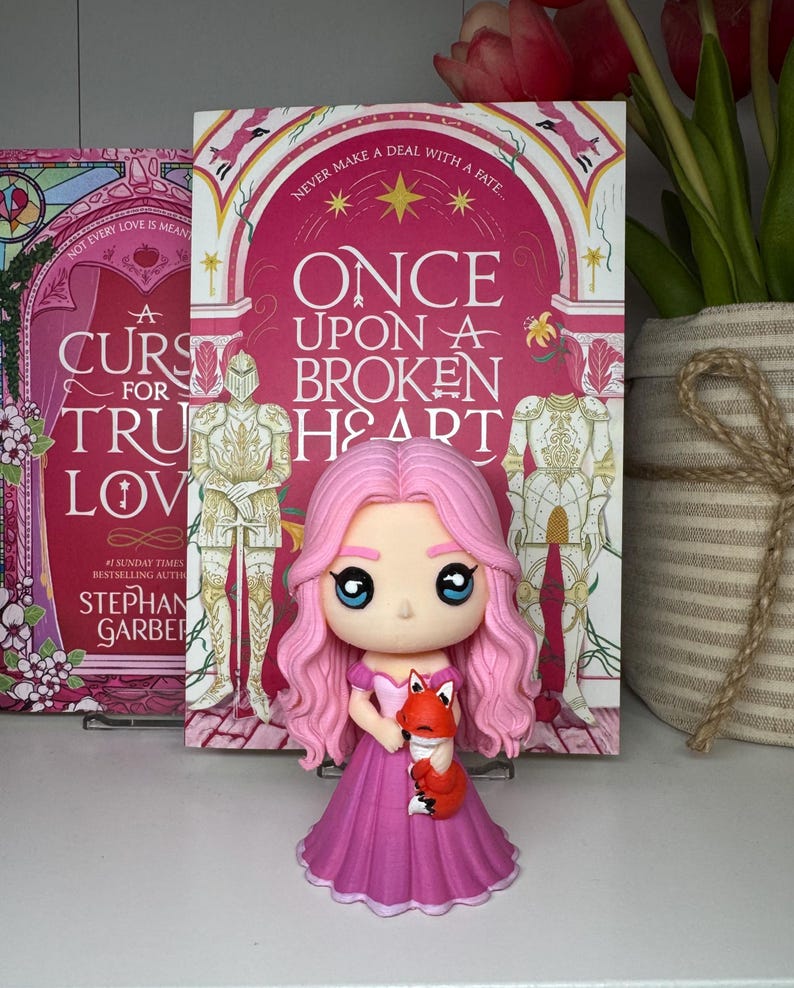 K&ouml;nnte beinhalten: Eine rosa Prinzessinnenfigur, die einen kleinen orangefarbenen Fuchs h&auml;lt, vor einem Buch mit dem Titel "Once Upon a Broken Heart". Die Figur hat gro&szlig;e blaue Augen und rosa Haare. Das Buchcover ist rosa.