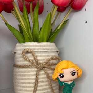 May include: A vase of red and pink tulips with green leaves, wrapped in beige fabric and tied with twine. A small figurine of a woman in a green dress is placed beside the vase.