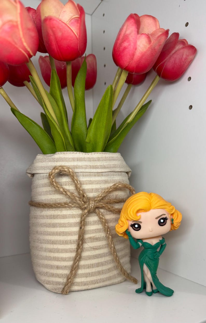 May include: A vase of red and pink tulips with green leaves, wrapped in beige fabric and tied with twine. A small figurine of a woman in a green dress is placed beside the vase.