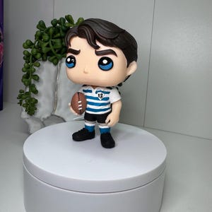 K&ouml;nnte beinhalten: Figur einer Person in Rugby-Uniform, die einen braunen Football h&auml;lt. Die Figur hat dunkles Haar, blaue Augen und tr&auml;gt ein wei&szlig;-blau gestreiftes Trikot mit der Nummer 13. Die Figur steht auf einer wei&szlig;en Plattform.