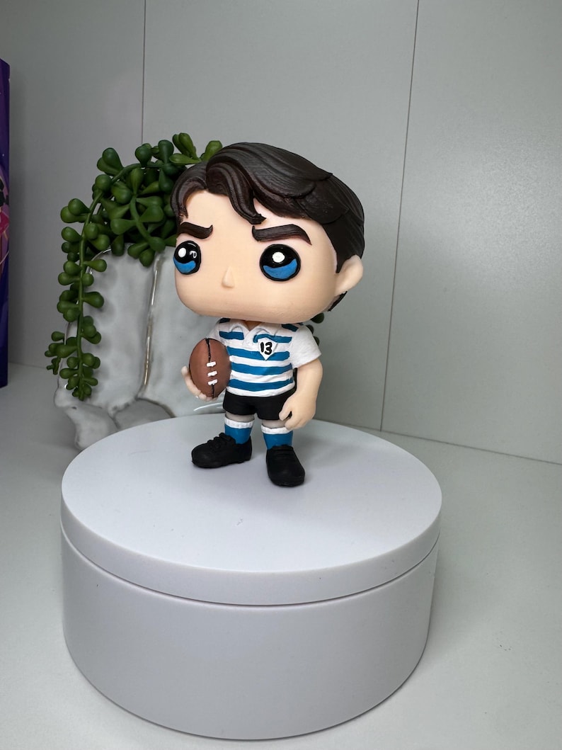 K&ouml;nnte beinhalten: Figur einer Person in Rugby-Uniform, die einen braunen Football h&auml;lt. Die Figur hat dunkles Haar, blaue Augen und tr&auml;gt ein wei&szlig;-blau gestreiftes Trikot mit der Nummer 13. Die Figur steht auf einer wei&szlig;en Plattform.
