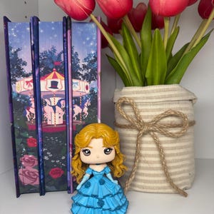 May include: A small figurine of a woman in a blue dress with blonde hair. The figurine is next to a vase of red tulips and three books with a carousel design. The vase is wrapped in beige fabric and tied with twine.