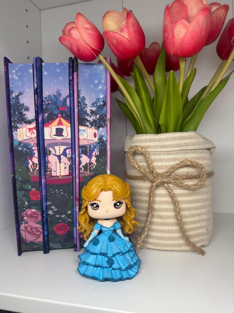 May include: A small figurine of a woman in a blue dress with blonde hair. The figurine is next to a vase of red tulips and three books with a carousel design. The vase is wrapped in beige fabric and tied with twine.