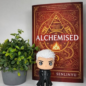 Może przedstawiać: Figurka Funko Pop z białymi włosami i czarnym płaszczem stoi przed książką zatytułowaną "ALCHEMISED" autorstwa Senlinyu. Książka ma czerwoną okładkę ze złotymi ilustracjami i tekstem. Mała roślina doniczkowa znajduje się po lewej stronie.