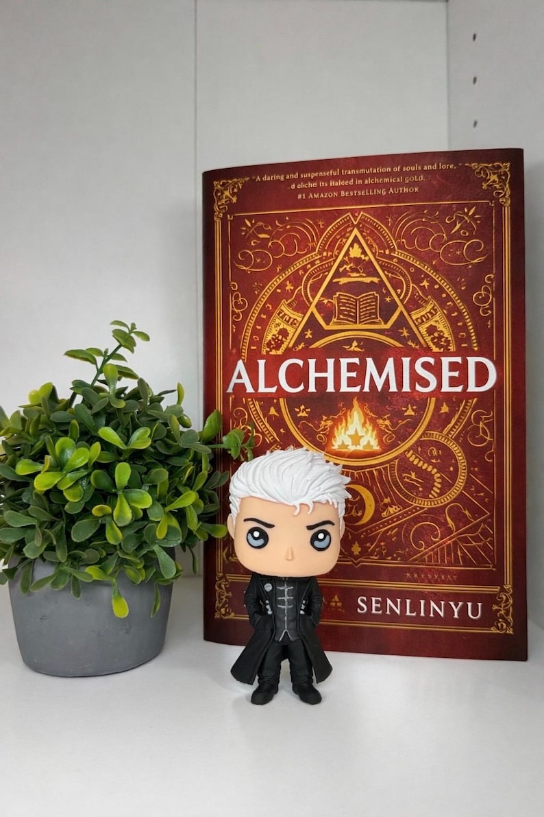 Może przedstawiać: Figurka Funko Pop z białymi włosami i czarnym płaszczem stoi przed książką zatytułowaną "ALCHEMISED" autorstwa Senlinyu. Książka ma czerwoną okładkę ze złotymi ilustracjami i tekstem. Mała roślina doniczkowa znajduje się po lewej stronie.