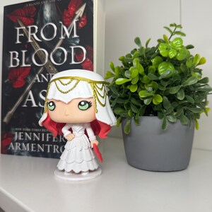 May include: A Funko Pop figure of a character from the book "From Blood and Ash" by Jennifer L. Armentrout. The figure is wearing a white dress and headdress, with red hair. A copy of the book and a small potted plant are also in the image.