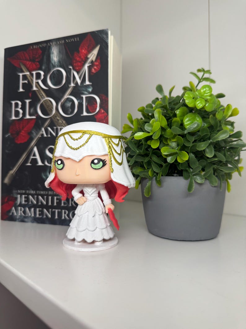 May include: A Funko Pop figure of a character from the book "From Blood and Ash" by Jennifer L. Armentrout. The figure is wearing a white dress and headdress, with red hair. A copy of the book and a small potted plant are also in the image.
