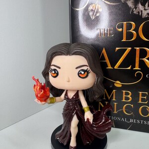 May include: A figurine of a woman in a burgundy dress holding a flame. The figure has dark hair and is on a black base. The book behind the figure has the title "The Bone Season" and the author's name.