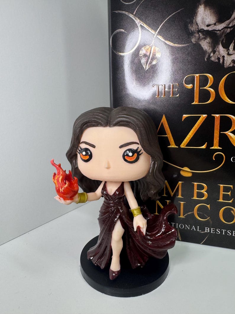 May include: A figurine of a woman in a burgundy dress holding a flame. The figure has dark hair and is on a black base. The book behind the figure has the title "The Bone Season" and the author's name.