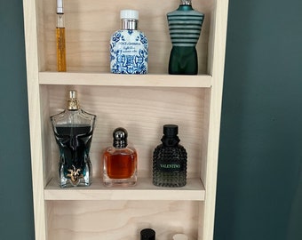 May include: A wooden shelf unit with three shelves, displaying a collection of perfume bottles. The bottles are various colors and shapes, including clear glass, blue and white porcelain, and black glass with a silver cap. The shelf unit is mounted on a dark green wall.