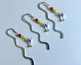 May include: Three silver-toned metal bookmarks with a wavy design. Each bookmark features a dangling charm shaped like a martini glass, accented with one small red bead and one oval-shaped yellow bead attached to the top curve of the metal.