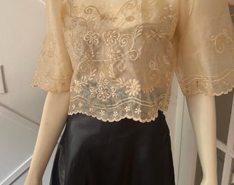May include: A beige, sheer top with floral embroidery and short, puffed sleeves. The top is paired with a black, satin skirt. The blouse features a scalloped hemline and is displayed on a mannequin.