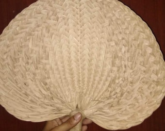 May include: A hand-held fan made of woven natural fibers. The fan has a heart-like shape with a looped handle. The weave pattern is visible, and the fan is a light beige color. The fan is held by a hand.