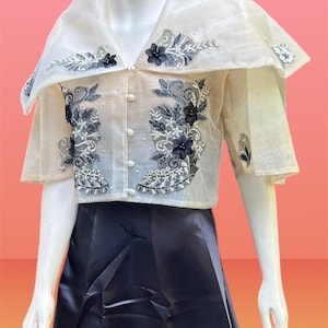 May include: A cream-coloured, sheer blouse with a high collar and short sleeves, decorated with intricate grey and black floral embroidery and pearl-like beads. The blouse is paired with a dark navy blue skirt, creating a formal ensemble.