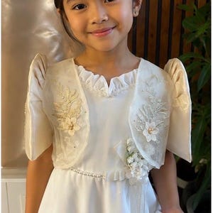 May include: A white dress featuring a ruffled neckline and a pearl-accented waistline. The dress is paired with a cream-colored bolero jacket that has structured butterfly sleeves, intricate floral embroidery, and small pearl embellishments.
