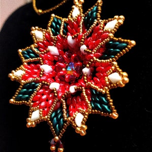 May include: A red, white, and green beaded pendant necklace with a gold chain. The pendant is shaped like a flower with a central crystal. The necklace has a tassel at the bottom. The text "Le B di Berenice" is visible below the pendant.