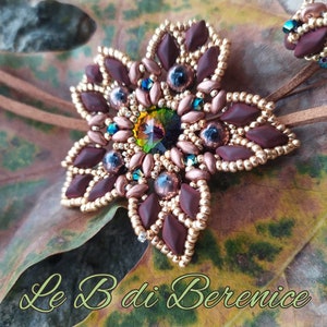 Può includere: Una collana con un ciondolo a forma di stella e un cordoncino in pelle marrone. Il ciondolo è fatto di perline marroni e dorate e presenta una grande pietra preziosa verde iridescente al centro. Il ciondolo è su una foglia marrone.