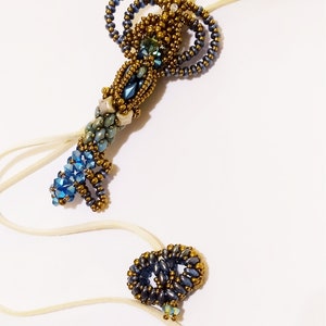 Può includere: Una collana con una chiave a forma di ciondolo in perle blu e dorate. La chiave è realizzata con piccole perle e ha un anello in cima per appenderla. La collana è realizzata con un cordoncino sottile e bianco.