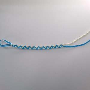 Puede incluir: Una pulsera de la amistad tejida a mano con un patrón geométrico repetitivo de ondas en hilo azul brillante y blanco roto. La pulsera incluye un extremo con bucle para el cierre y dos cordones trenzados largos que se extienden por el otro lado.
