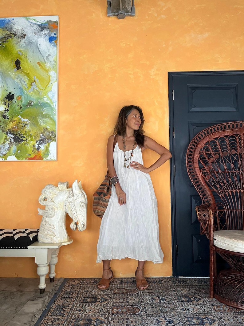 May include: A woman in a white sundress stands in a room with an orange wall. She wears brown sandals and carries a patterned bag. A carved white horse head sits on a white bench. A peacock chair and abstract art are also visible.
