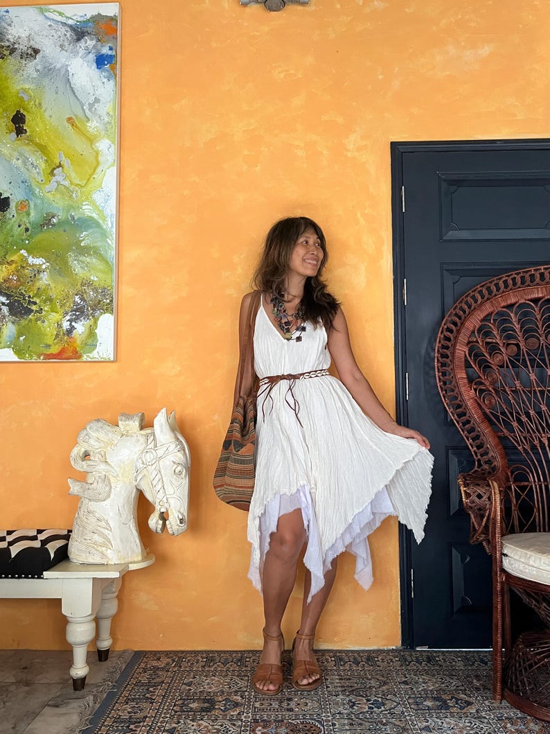 K&ouml;nnte beinhalten: Ein wei&szlig;es, asymmetrisches Kleid mit Spaghettitr&auml;gern und einem mehrlagigen Rock. Das Kleid wird mit braunen Sandalen und einer gewebten Tragetasche getragen. Ein brauner G&uuml;rtel betont die Taille. Der Hintergrund zeigt eine orangefarbene Wand und eine Pferdekopfskulptur.
