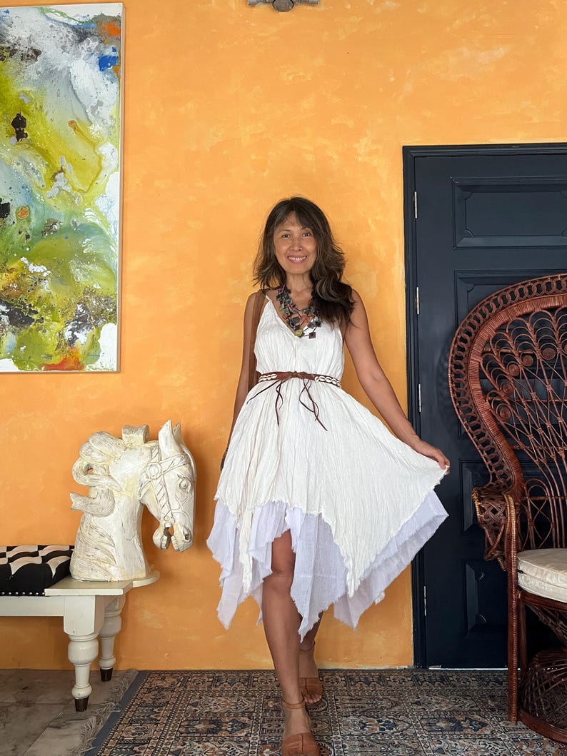 K&ouml;nnte beinhalten: Ein wei&szlig;es, asymmetrisches Kleid mit einem mehrschichtigen Rock und einem braunen G&uuml;rtel. Das Kleid hat ein flie&szlig;endes, strukturiertes Aussehen. Die Frau tr&auml;gt braune Sandalen und eine mehrreihige Halskette. Der Hintergrund zeigt eine orangefarbene Wand, eine dunkelblaue T&uuml;r und einen dekorativen Stuhl.