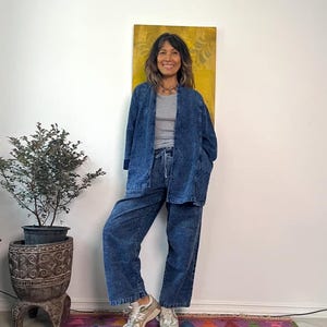 May include: A woman wearing a denim jacket, a gray top, and matching striped denim pants. The outfit is paired with gray sneakers. A colorful rug and a potted plant are in the background. The jacket has large pockets.