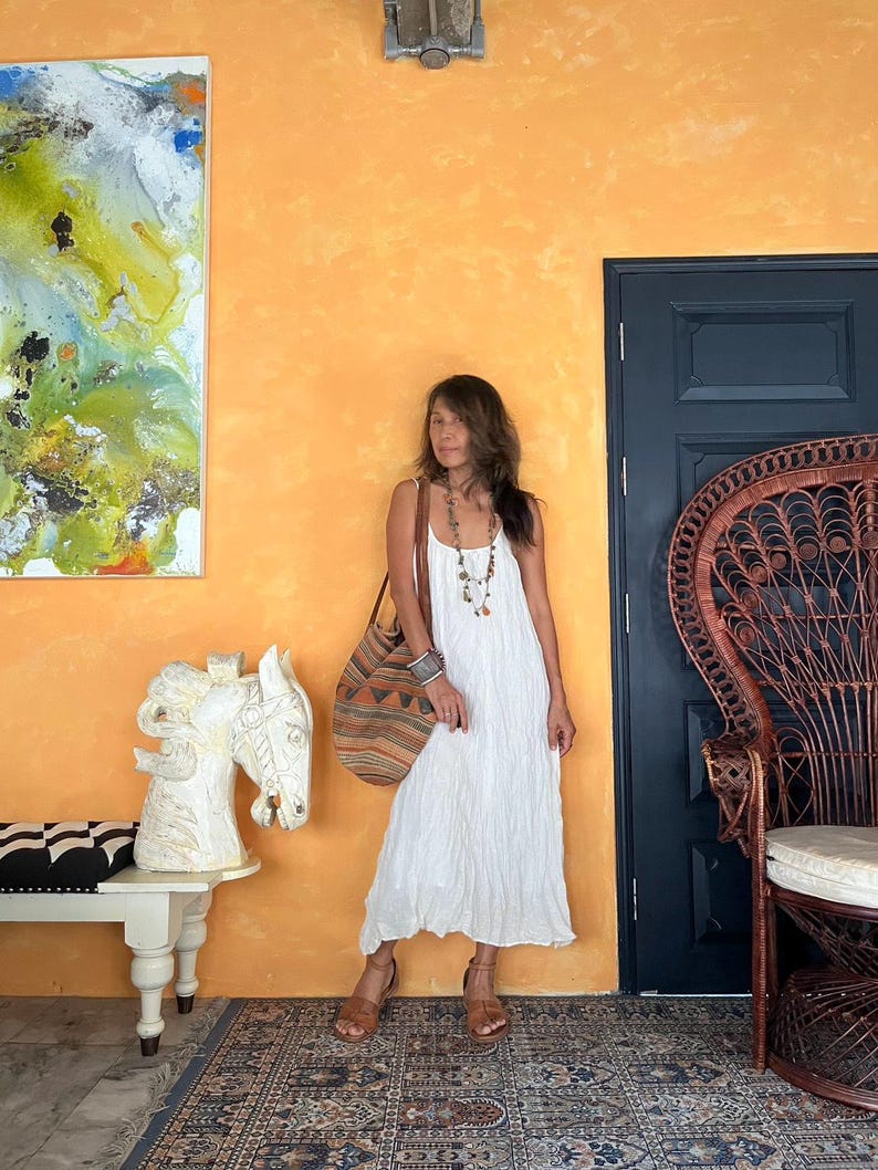 May include: A woman wearing a white maxi dress and brown sandals stands in front of an orange wall. She carries a large, patterned tote bag and wears a beaded necklace. A white horse sculpture and a peacock chair are also visible.