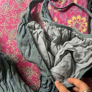 May include: Close-up of a teal-colored garment with a gathered neckline and tie straps. The fabric appears soft and flowing, with visible texture. The garment is set against a pink floral patterned background.