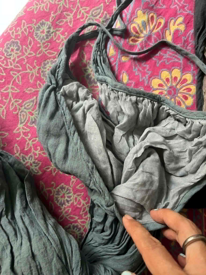 May include: Close-up of a teal-colored garment with a gathered neckline and tie straps. The fabric appears soft and flowing, with visible texture. The garment is set against a pink floral patterned background.