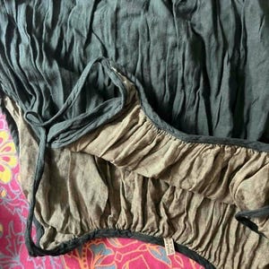 May include: Close-up of a dark teal-colored garment with a ruffled edge and a contrasting tan lining. The fabric appears to be lightweight and textured, with a drawstring detail. The garment is set against a vibrant pink patterned background.