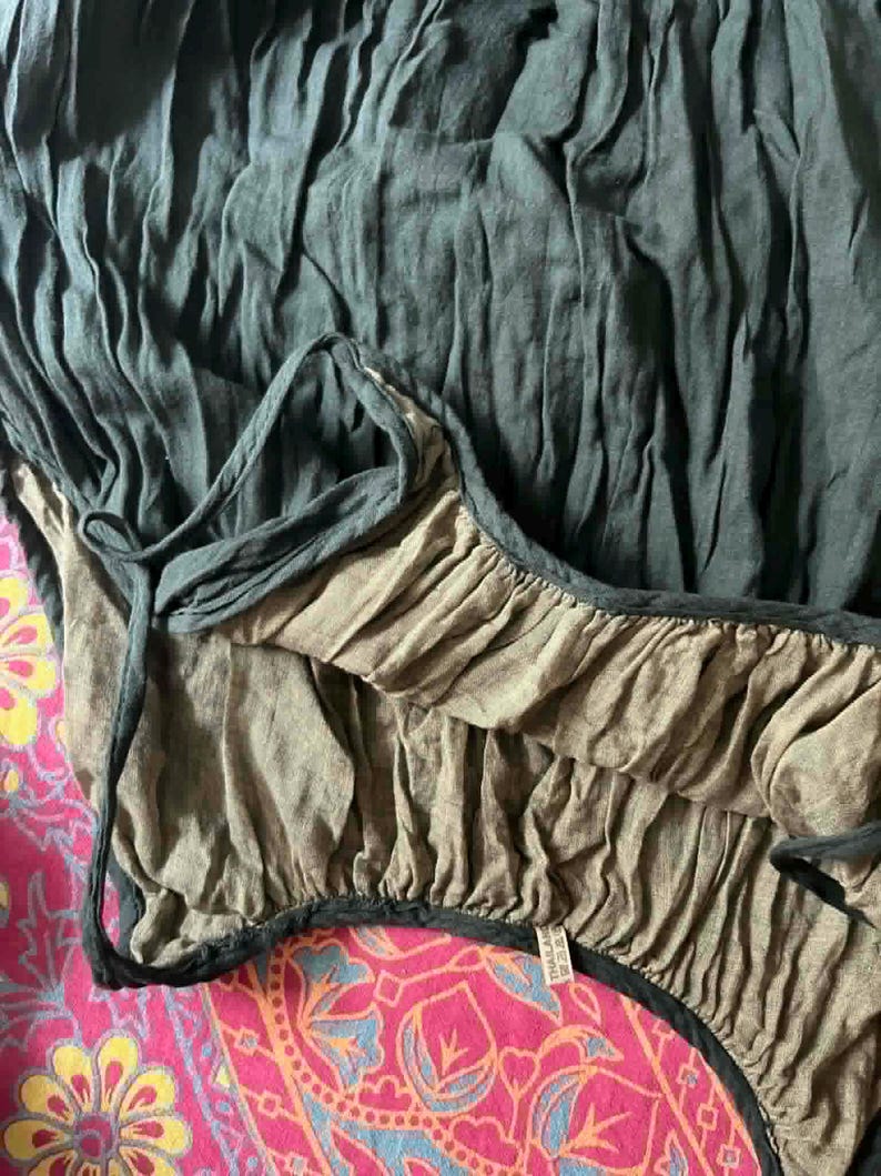May include: Close-up of a dark teal-colored garment with a ruffled edge and a contrasting tan lining. The fabric appears to be lightweight and textured, with a drawstring detail. The garment is set against a vibrant pink patterned background.