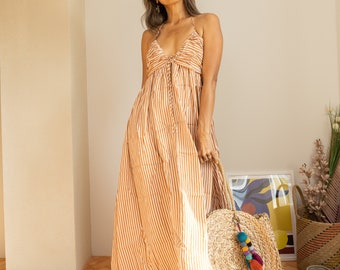 Könnte beinhalten: Eine Frau in einem langen, orange-weiß gestreiften Sommerkleid mit V-Ausschnitt und Spaghettiträgern. Sie hält eine große Stroh-Tragetasche mit einem bunten Quastenanhänger. Das Kleid ist bodenlang, und sie trägt Sandalen.