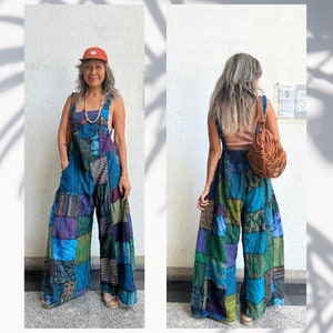 May include: A woman is wearing a patchwork denim overall jumpsuit with a brown leather bag. The jumpsuit has a variety of colors and patterns, including blue, green, purple, and black. The woman is standing in front of a white wall with a shadow of a plant on it.