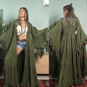 May include: Olive green maxi dress with long, flowing sleeves and tiered skirt. The dress features ruffled edges and a relaxed fit. The model is wearing a white crochet top and denim shorts underneath the dress.