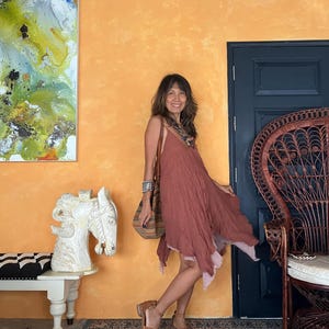 K&ouml;nnte beinhalten: Ein flie&szlig;endes, staubrosa Kleid mit einem geschichteten Saum. Das Kleid wird mit braunen Sandalen und einer gestreiften Umh&auml;ngetasche kombiniert. Der Hintergrund zeigt eine orangefarbene Wand, eine dunkelblaue T&uuml;r und einen dekorativen Pfauenstuhl.