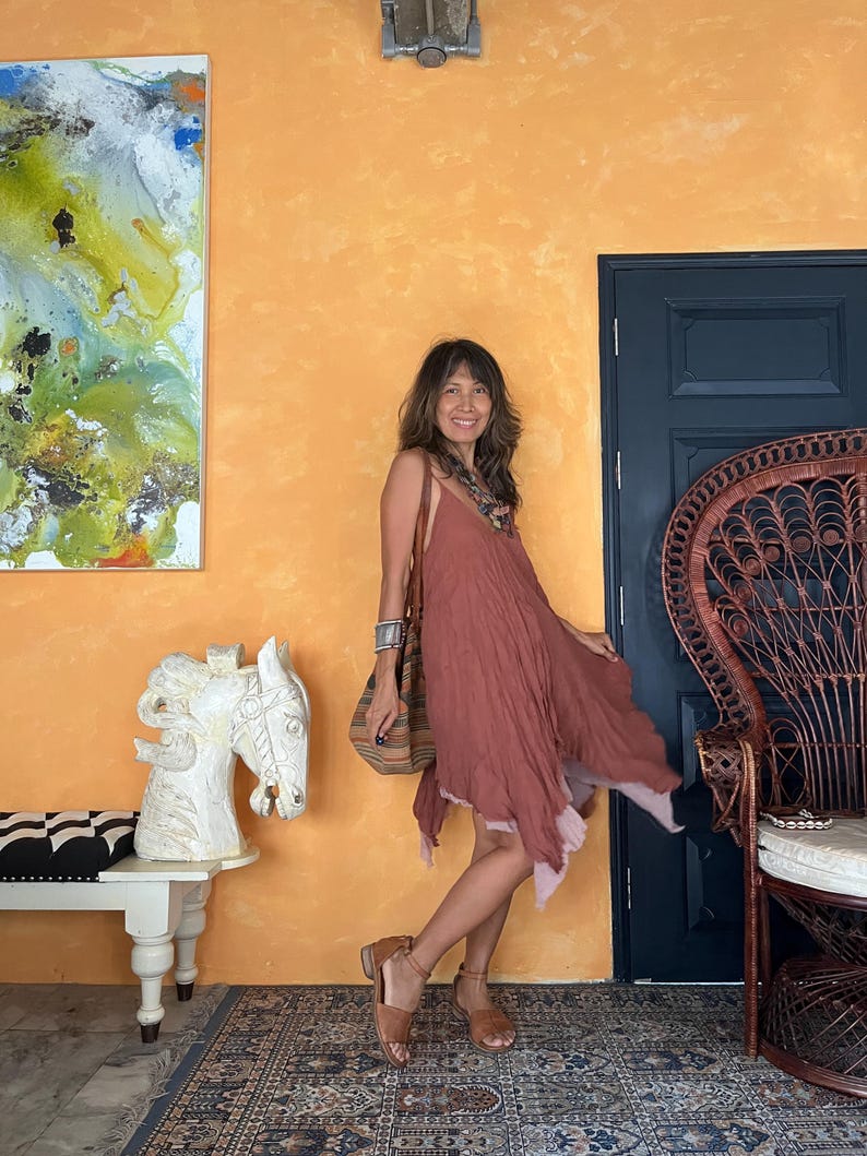 K&ouml;nnte beinhalten: Ein flie&szlig;endes, staubrosa Kleid mit einem geschichteten Saum. Das Kleid wird mit braunen Sandalen und einer gestreiften Umh&auml;ngetasche kombiniert. Der Hintergrund zeigt eine orangefarbene Wand, eine dunkelblaue T&uuml;r und einen dekorativen Pfauenstuhl.