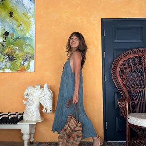 May include: A woman in a flowing teal dress and brown sandals carries a striped tote bag with orange and brown accents. The background features an orange wall, a dark blue door, and a decorative chair.