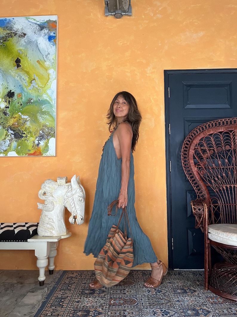 May include: A woman in a flowing teal dress and brown sandals carries a striped tote bag with orange and brown accents. The background features an orange wall, a dark blue door, and a decorative chair.